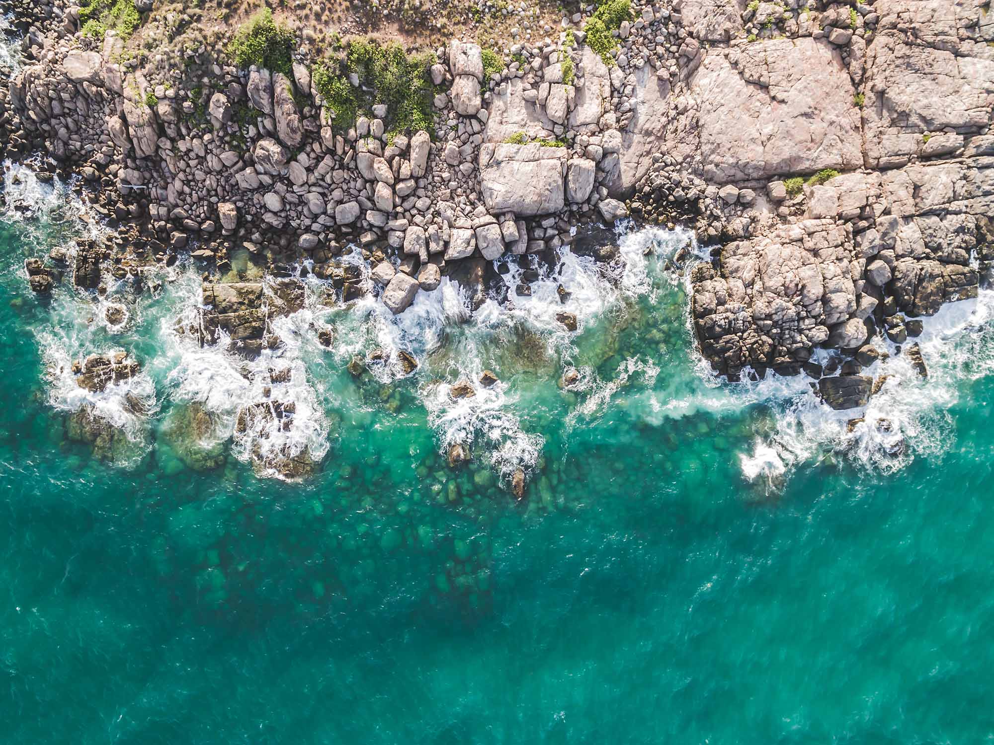 Top down aerial photo of a rocky coastline with turquoise water crashing up against the granite rocks