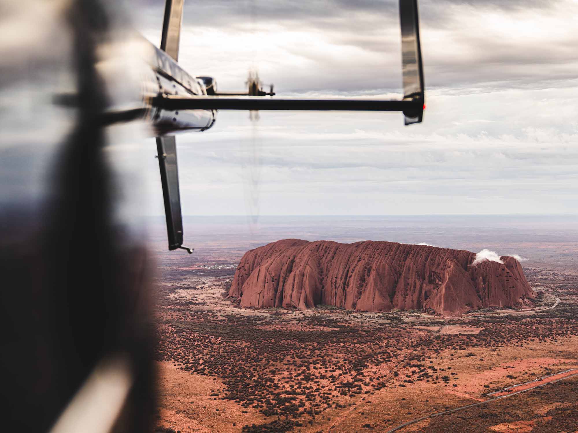 Handheld aerial photo on an overcast day, looking back towards the tail of a helicopter flying over a full view of Uluru which you can see in the background that also has small clouds sitting on top of the rock itself